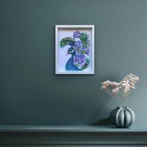 Op de afbeelding: Een ingelijst papieren kunstwerk dat een pauw met blauwe en groene veren en een paarse bloemenboeket voorstelt. Het kunstwerk is op een witte achtergrond en is ingelijst in een wit frame.