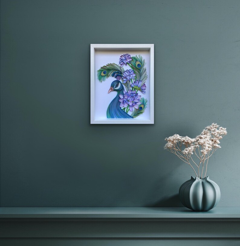 Op de afbeelding: Een ingelijst papieren kunstwerk dat een pauw met blauwe en groene veren en een paarse bloemenboeket voorstelt. Het kunstwerk is op een witte achtergrond en is ingelijst in een wit frame.