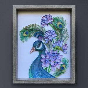 Op de afbeelding: Een ingelijst papier-quilling kunstwerk met een pauw met blauwe en groene veren en een paarse bloemenboeket. Het kunstwerk is op een witte achtergrond en ingelijst in een grijs houten frame.