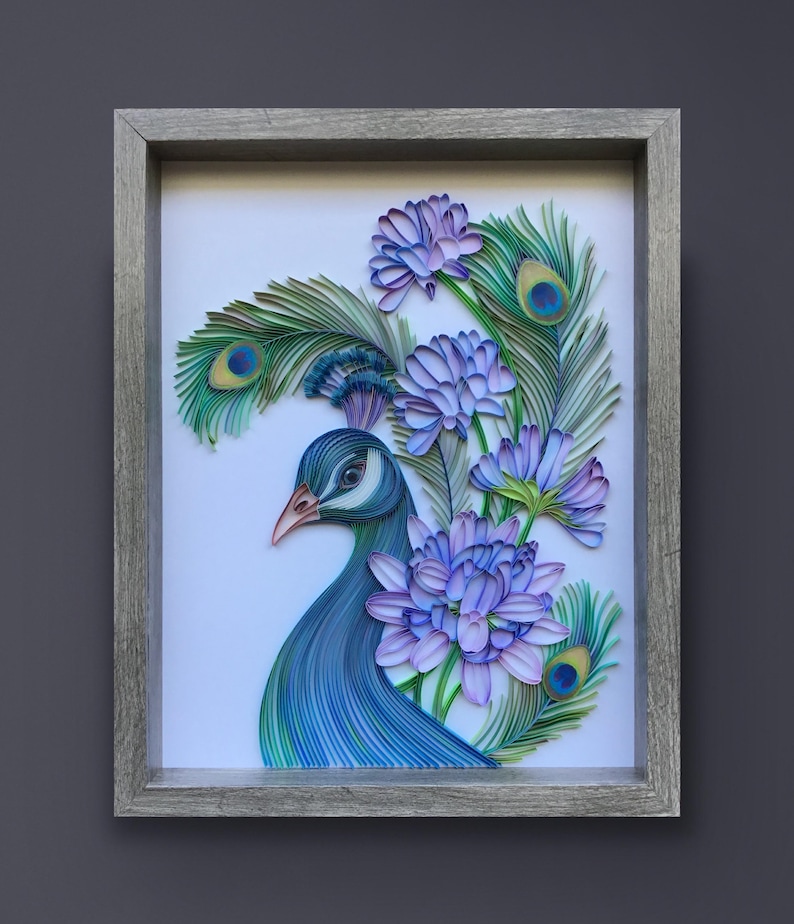 Op de afbeelding: Een ingelijst papier-quilling kunstwerk met een pauw met blauwe en groene veren en een paarse bloemenboeket. Het kunstwerk is op een witte achtergrond en ingelijst in een grijs houten frame.