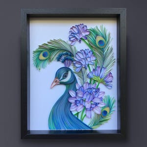 Op de afbeelding: Een ingelijst kunstwerk van papier met een pauw met blauwe en groene veren en een paarse bloemenboeket. De kop van de pauw is naar de zijkant gedraaid, en zijn oog is zichtbaar. De bloemen zijn rond de nek en staart van de pauw gerangschikt.