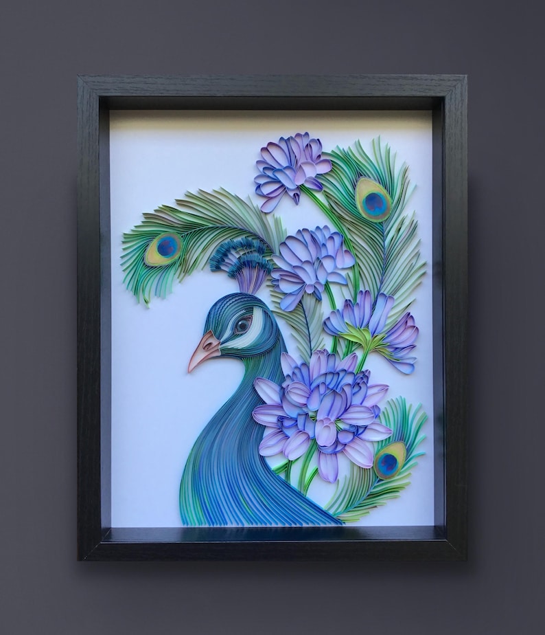 Op de afbeelding: Een ingelijst kunstwerk van papier met een pauw met blauwe en groene veren en een paarse bloemenboeket. De kop van de pauw is naar de zijkant gedraaid, en zijn oog is zichtbaar. De bloemen zijn rond de nek en staart van de pauw gerangschikt.
