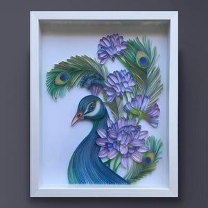 Op de afbeelding: Een ingelijst papieren kunstwerk met een pauw met blauwe en groene veren en paarse bloemen. De kop van de pauw is naar de zijkant gedraaid, en zijn oog is zichtbaar. De bloemen zijn rond de kop en nek van de pauw gerangschikt.