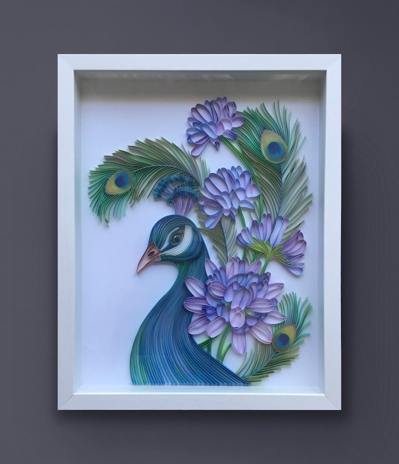 Op de afbeelding: Een ingelijst papieren kunstwerk met een pauw met blauwe en groene veren en paarse bloemen. De kop van de pauw is naar de zijkant gedraaid, en zijn oog is zichtbaar. De bloemen zijn rond de kop en nek van de pauw gerangschikt.