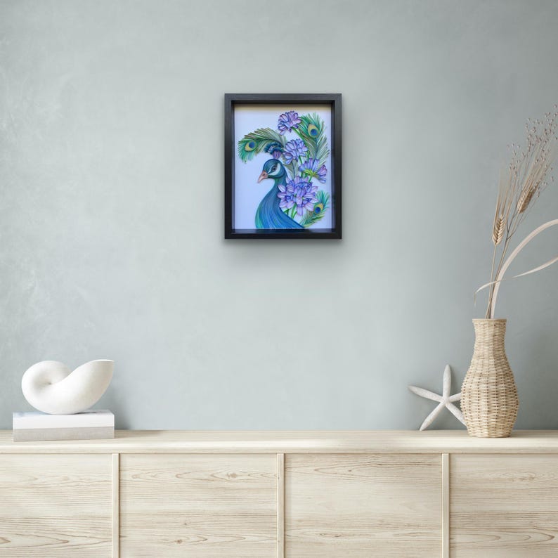 Op de afbeelding: Een ingelijste illustratie van een pauw met blauwe en groene veren en paarse bloemen. De pauw kijkt naar links in de afbeelding. De lijst is zwart.