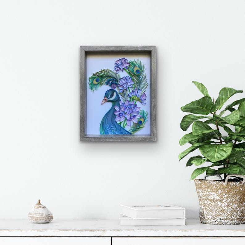 Op de afbeelding: Een ingelijst papier-quilling kunstwerk met een pauw met blauwe en groene veren en paarse bloemen.