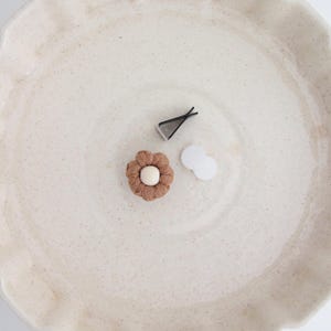 May include: A cream-coloured, speckled ceramic plate with a scalloped edge. The plate holds a brown flower-shaped hair clip, a silver hair clip, and two small white circles. The items are arranged in the centre of the plate.