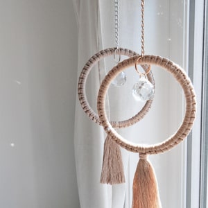Pode incluir: Dois aros de macramé bege com borlas penduradas em uma janela. Cada aro tem um prisma de cristal pendurado.
