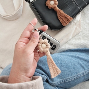Puede incluir: Un llavero de macramé beige con forma de flor y borla, sujeto a un mando de coche. Un segundo llavero de macramé con forma de flor en color marrón oscuro está sujeto a un bolso negro. Los llaveros son hechos a mano y decorativos.