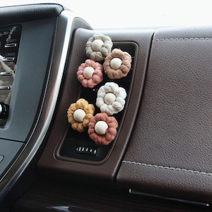May include: Six crocheted flower car air freshener clips in various colours, including beige, brown, peach, and green. The flowers are attached to a car air vent.