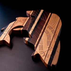 May include: Two wooden cribbage boards shaped like fish. The boards are crafted from various wood tones, including light, medium, and dark browns, with black stripes. Each board has drilled holes for pegs.