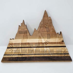 May include: Wooden cribbage board shaped like a mountain range. The board has a variety of wood tones and the words "SCORE" and "FINISH". It features multiple rows of pegging holes and a dark brown stripe across the board.