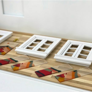 K&ouml;nnte beinhalten: Drei wei&szlig;e Miniaturfenster mit Holzrahmen und Glasscheiben. Vier rechteckige St&uuml;cke aus klarem Acryl mit einem bunten abstrakten Design liegen auf einem Holzboden.