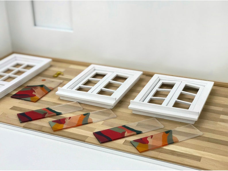 K&ouml;nnte beinhalten: Drei wei&szlig;e Miniaturfenster mit Holzrahmen und Glasscheiben. Vier rechteckige St&uuml;cke aus klarem Acryl mit einem bunten abstrakten Design liegen auf einem Holzboden.