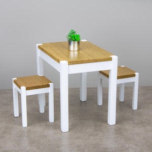 Peut inclure: Une table blanche avec un plateau en bois et deux tabourets blancs. La table est dressée avec une petite plante en pot.