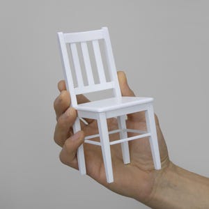 Peut inclure: Une chaise en bois blanc avec un design simple. La chaise est tenue dans la main d'une personne.