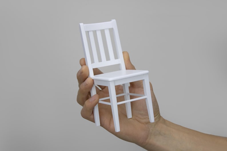 Peut inclure: Une chaise en bois blanc avec un design simple. La chaise est tenue dans la main d'une personne.