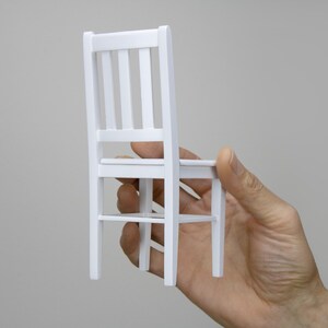 Peut inclure: Une chaise en bois blanc avec un design simple. La chaise est tenue dans une main, montrant le dessous de l'assise et les pieds.