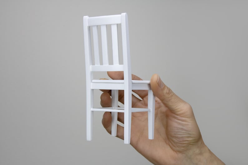 Peut inclure: Une chaise en bois blanc avec un design simple. La chaise est tenue dans une main, montrant le dessous de l'assise et les pieds.