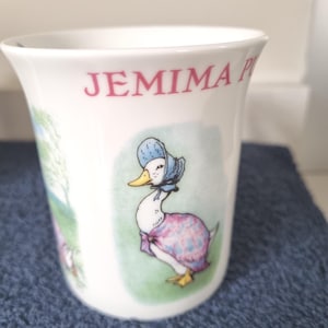 Op de afbeelding: Een witte keramische mok met een kleurrijke illustratie van een eend die een blauwe muts en een roze sjaal met patroon draagt. De tekst JEMIMA P is in rode letters bij de bovenrand gedrukt. De mok heeft een gladde, uitlopende rand.