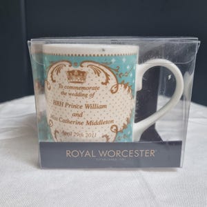 Pode incluir: Uma caneca de cerâmica branca com um padrão decorativo em turquesa e dourado, apresentando o texto To commemorate the wedding of HRH Prince William and Miss Catherine Middleton April 29th 2011, numa caixa de plástico transparente Royal Worcester.