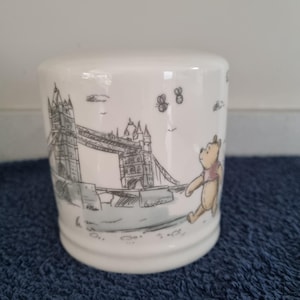 May include: A white ceramic mug featuring a black line drawing of Tower Bridge in London. The illustration includes a small, light brown bear character walking near the bridge, accompanied by two small flying bees in the sky above the structure.