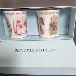 May include: Two white ceramic mugs featuring Beatrix Potter illustrations, housed in a light blue gift box labeled BEATRIX POTTER. The left mug shows Mrs. Tiggy-Winkle, and the right mug depicts Benjamin Bunny with green text above the character art.