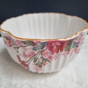 May include: A white ceramic bowl with a scalloped rim and a gold-painted edge. The exterior features a floral pattern with pink and red blossoms, green leaves, and brown stems. The bowl has a vertical ribbed texture around the entire outer surface.