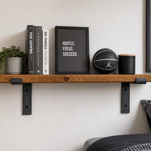 May include: A rustic wooden wall shelf supported by two black metal brackets. The shelf holds a potted green plant, three books, a black framed sign with the text HUSTLE. FOCUS. SUCCEED., a black basketball, and a black cylindrical container.