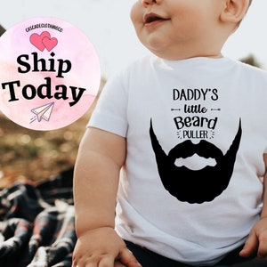 May include: A white baby onesie with black text that reads "Daddy's little Beard Puller" and a black silhouette of a beard.