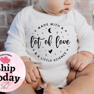 May include: White baby onesie with long sleeves. The onesie has a circular graphic with the text "Made with a lot of love & a little science". The design includes a crescent moon, stars, and a heart.
