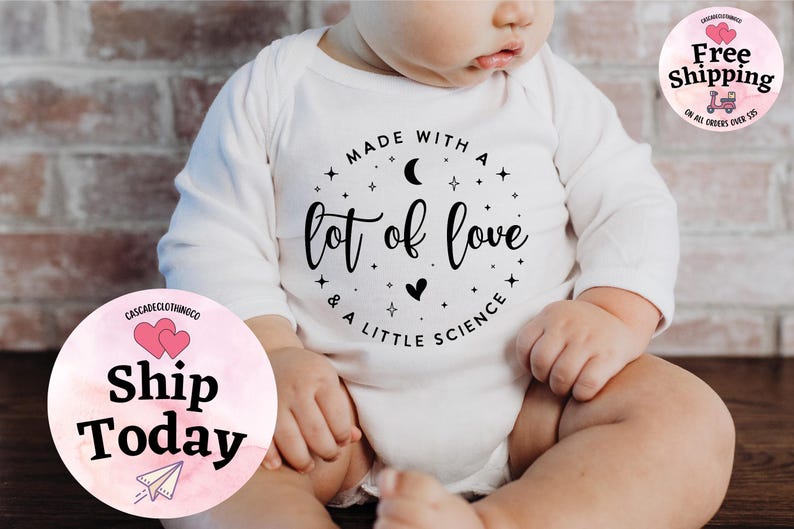 May include: White baby onesie with long sleeves. The onesie has a circular graphic with the text "Made with a lot of love & a little science". The design includes a crescent moon, stars, and a heart.