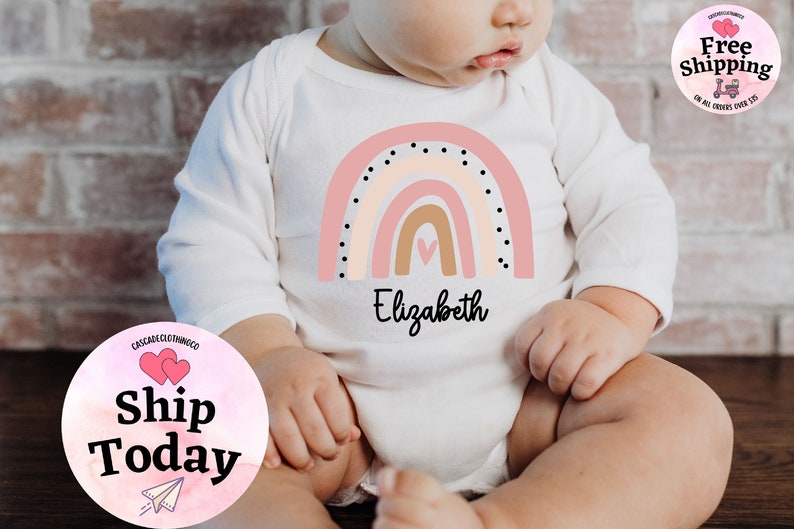 May include: A white long-sleeved onesie with a pink, beige, and brown rainbow design. The rainbow has black polka dots and a small pink heart in the center. The onesie is personalized with the name "Elizabeth".