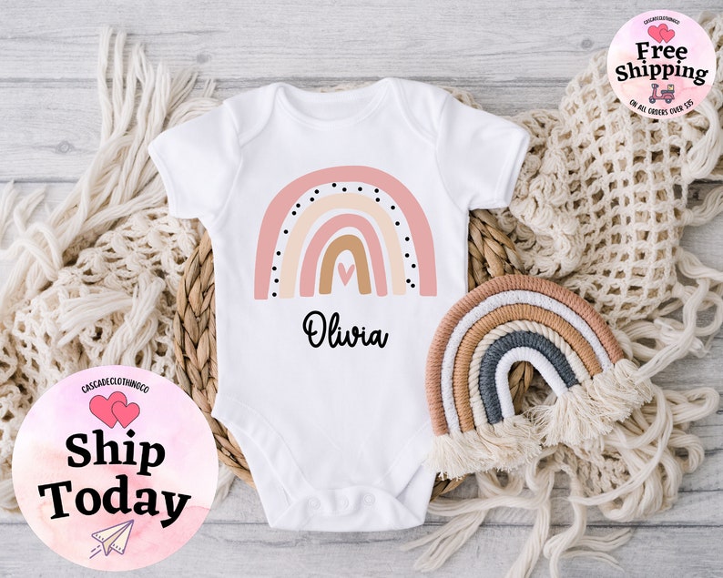 May include: White baby onesie with a pink, orange, and brown rainbow design and black polka dots. The onesie has the name "Olivia" printed below the rainbow.