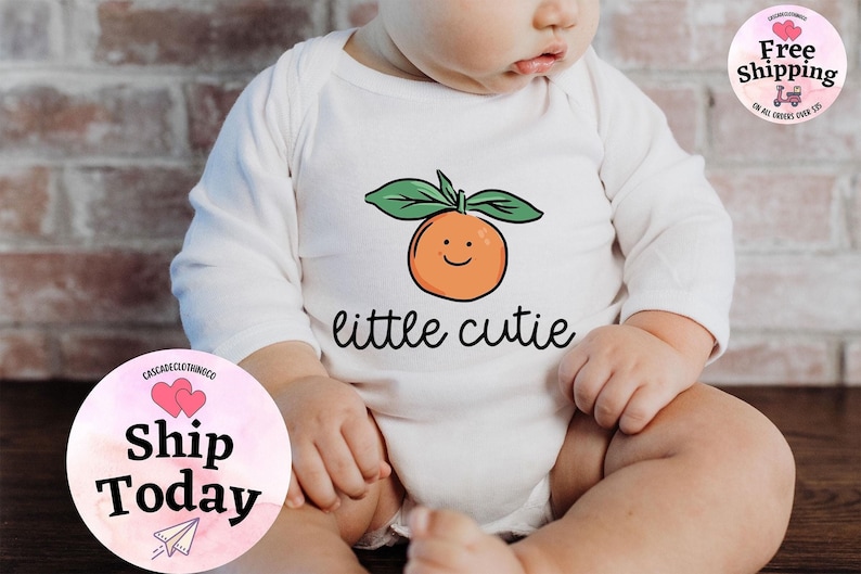 May include: White long-sleeve baby onesie with a cartoon orange and the text "little cutie".