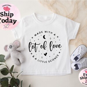 May include: White toddler t-shirt with the text "Made with a lot of love & a little science" in a circular design. The shirt is paired with white sneakers and a gray koala plush toy. The image also includes a "Ship Today" and "Free Shipping" graphic.
