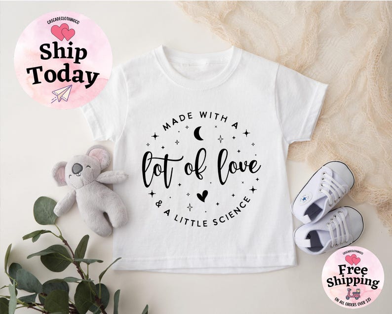 May include: White toddler t-shirt with the text "Made with a lot of love & a little science" in a circular design. The shirt is paired with white sneakers and a gray koala plush toy. The image also includes a "Ship Today" and "Free Shipping" graphic.