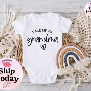 May include: White baby onesie with black text that reads "Pass me to grandma" with a small heart. The onesie is on a white background with a woven basket and a rainbow.