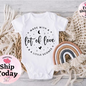 May include: White baby onesie with the text "Made with a lot of love & a little science" in a black script. A small, decorative rainbow and a woven basket are also in the image.