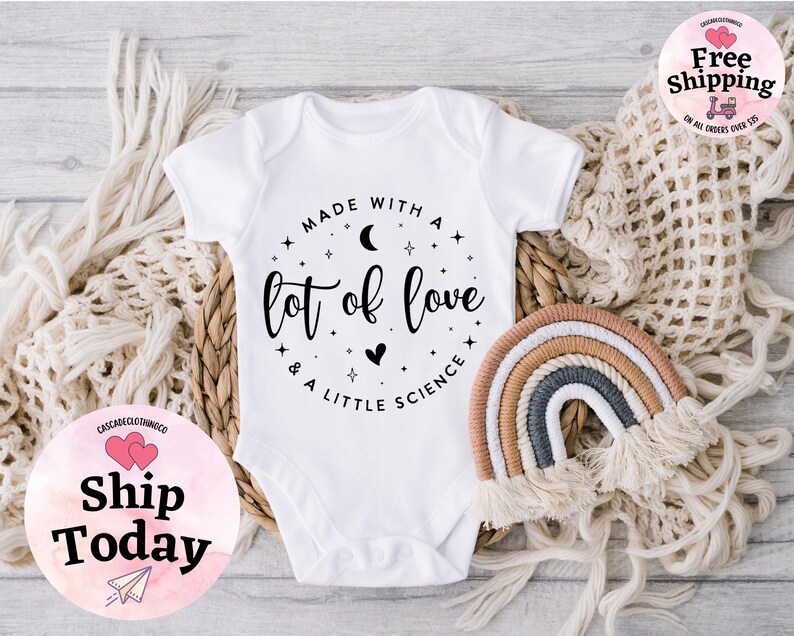 May include: White baby onesie with the text "Made with a lot of love & a little science" in a black script. A small, decorative rainbow and a woven basket are also in the image.