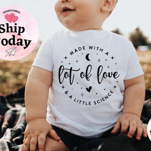 May include: White baby t-shirt with the text "Made with a lot of love & a little science" in black script. The design includes stars, a crescent moon, and a heart. The shirt is short-sleeved.