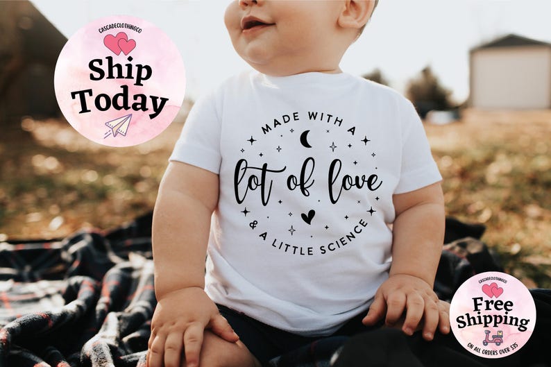 May include: White baby t-shirt with the text "Made with a lot of love & a little science" in black script. The design includes stars, a crescent moon, and a heart. The shirt is short-sleeved.