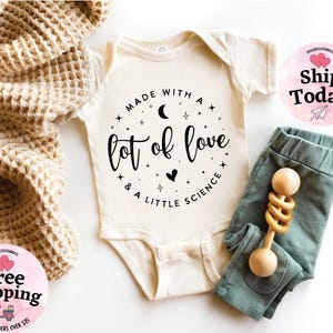 May include: Cream-colored baby bodysuit with the text "MADE WITH A lot of love & A LITTLE SCIENCE" in black. Paired with teal-colored baby pants and a wooden rattle. A waffle-knit blanket is draped on the side.