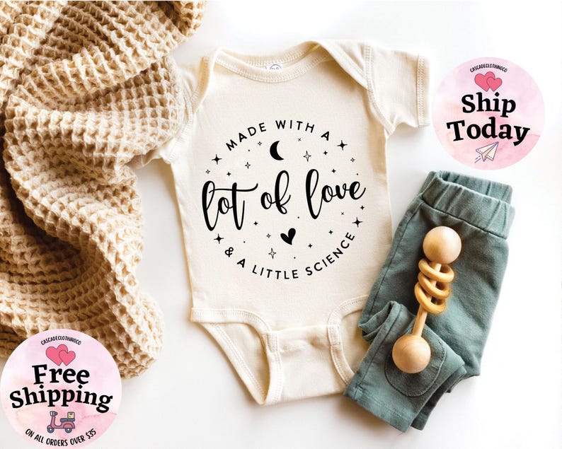 May include: Cream-colored baby bodysuit with the text "MADE WITH A lot of love & A LITTLE SCIENCE" in black. Paired with teal-colored baby pants and a wooden rattle. A waffle-knit blanket is draped on the side.