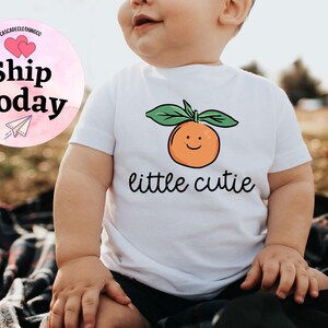 May include: White baby onesie with a smiling orange graphic and the text "little cutie".