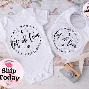 May include: White baby onesie and bib set. Both items feature the text "Made with a lot of love & a little science" in a circular design with stars, moons, and hearts. The set is designed for infants.