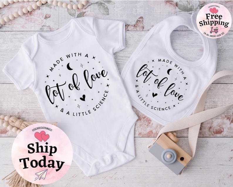 May include: White baby onesie and bib set. Both items feature the text "Made with a lot of love & a little science" in a circular design with stars, moons, and hearts. The set is designed for infants.