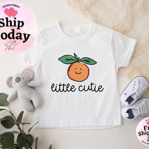 May include: White baby onesie with a smiling orange and the text "little cutie" printed on it.