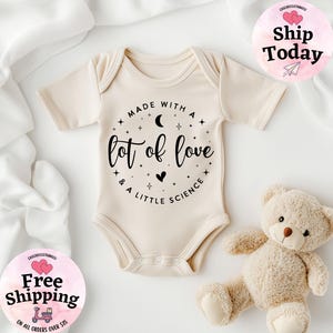 May include: Cream-colored baby onesie with the text "Made with a lot of love & a little science" in black script. The onesie has short sleeves and snaps at the bottom. A teddy bear is next to the onesie.