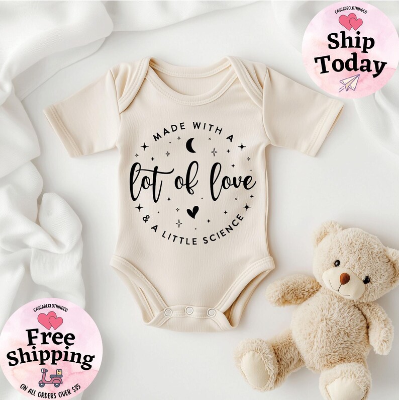May include: Cream-colored baby onesie with the text "Made with a lot of love & a little science" in black script. The onesie has short sleeves and snaps at the bottom. A teddy bear is next to the onesie.
